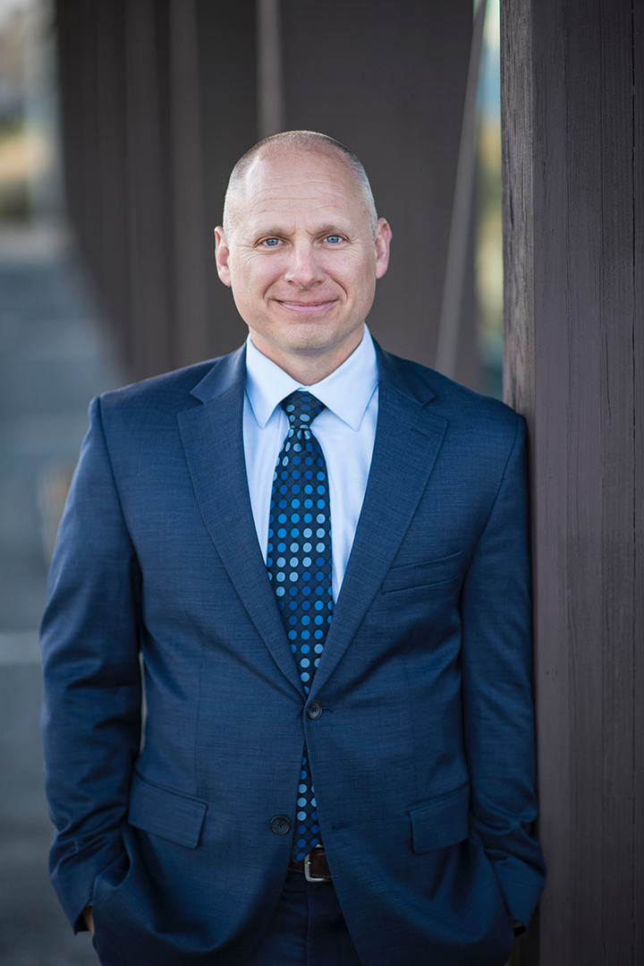 man with short hair, blue tailored suit and blue patterned tie and collared shirt with abstract outdoor background