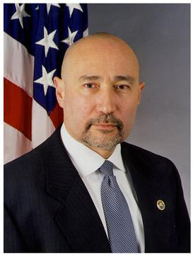 Headshot of John Perren in dark suit, white shirt, tie with U.S. flag behind him