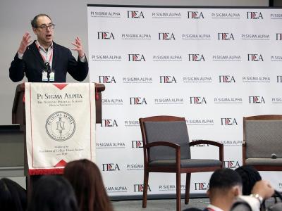 Matt Dallek speaking at a podium, next to a Pi Sigma Alpha Student Conference step-and-repeat