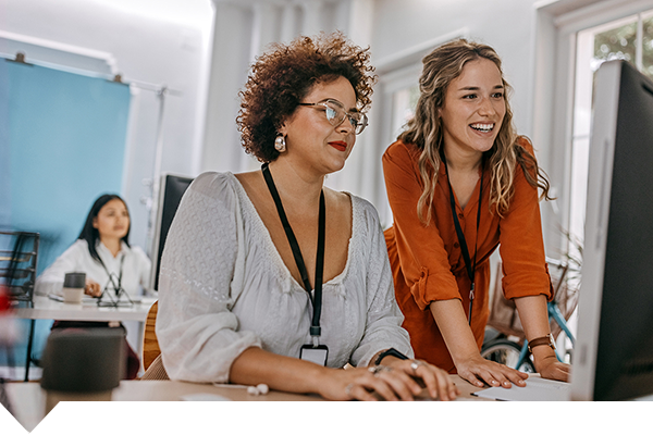 two women look at a computer screen together with another person in the background in a classroom environment