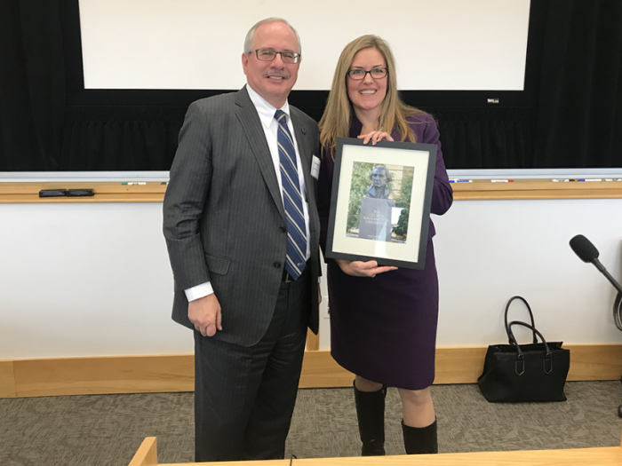 U.S. Rep. Wexton, professional attire, long blond hair, stands to the right of GW Pres. LeBlanc in suit. She is holding a photo.