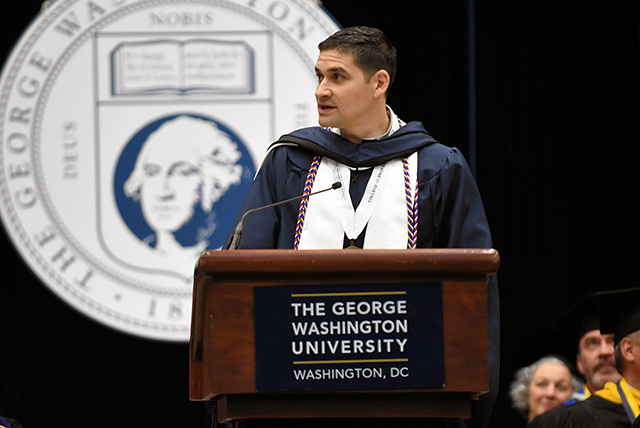 student speaker at podium in cap and gown with GW seal behind