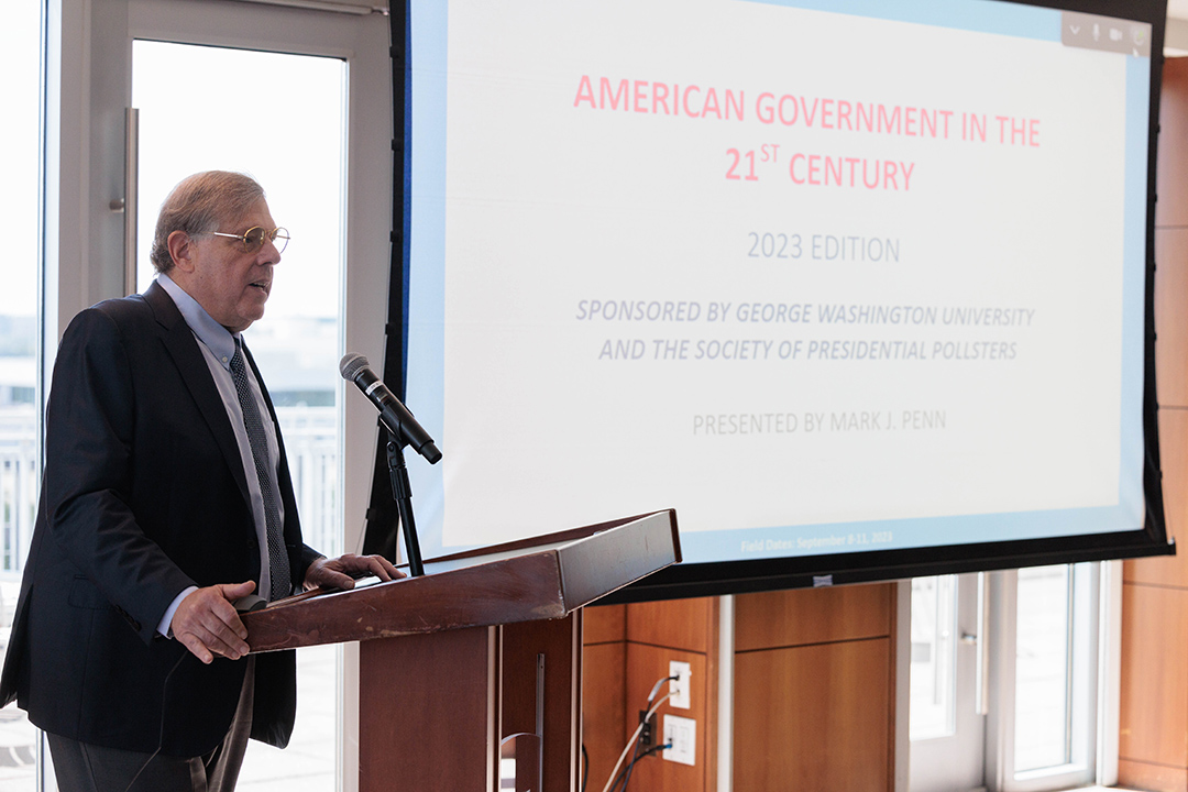 Mark Penn standing and speaking in front of a projector screen that displays the Presidential Pollsters title slide