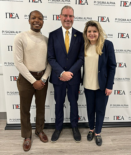  Professor Todd Belt, Nick Key and Stami Williams all standing and smiling in front of a step-and-repeat
