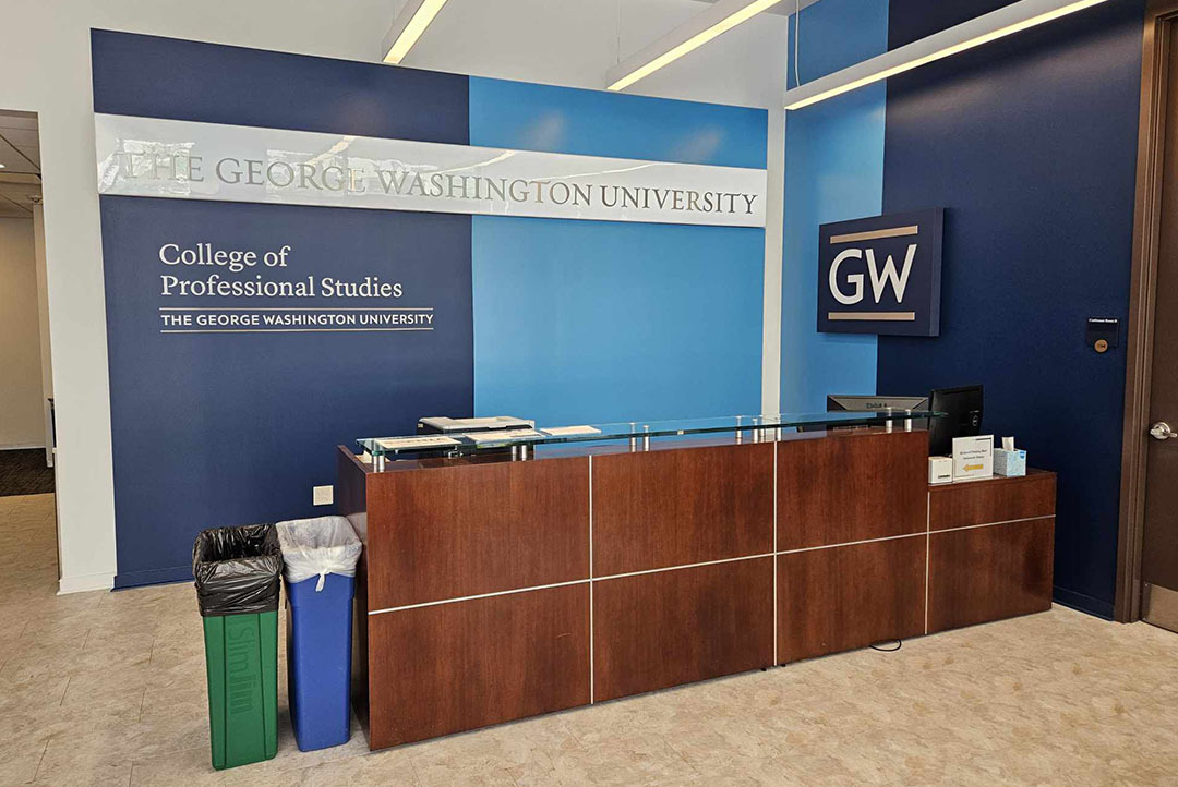 Reception area with a blue wall behind it. The left of the wall is dark blue and has the College of Professional studies logo on it. The other half of the wall is a light, bright blue with no text on it.