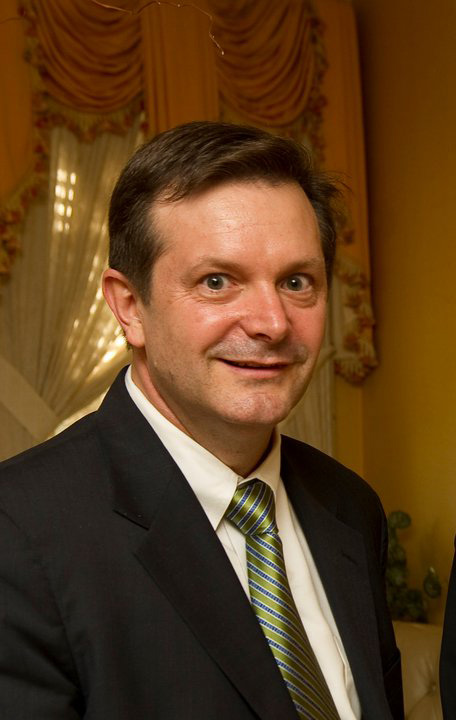 Dean Smith in dark suit, white shirt, shiny moss green tie and brown hair with curtains in the background