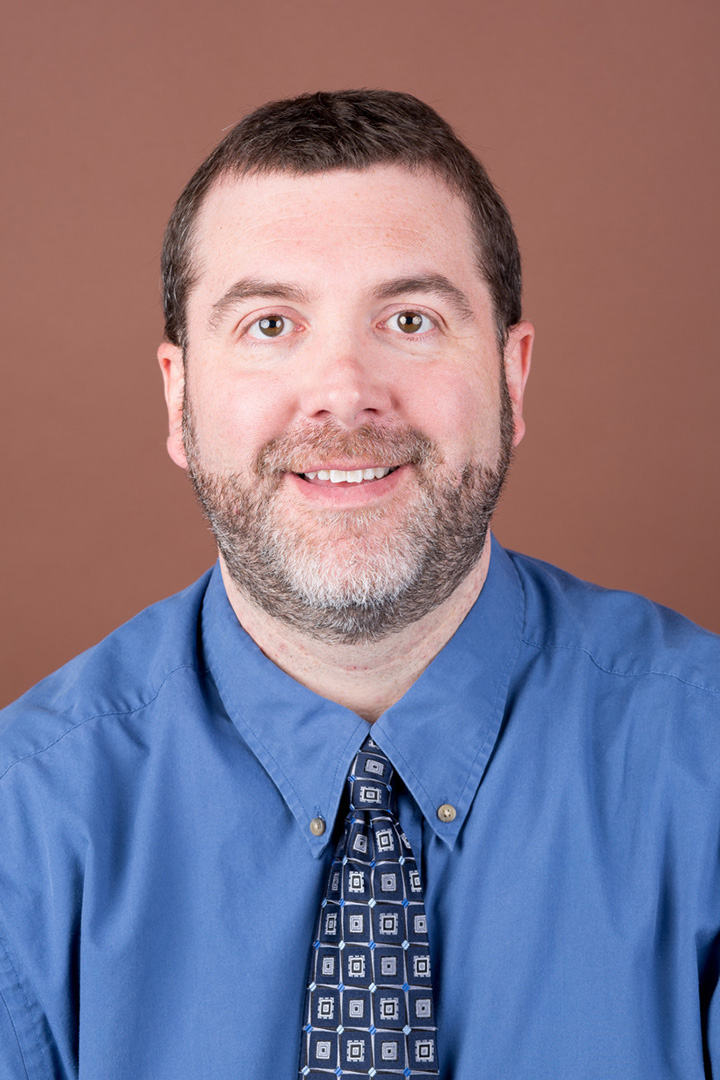 Mike Kalyan in blue collared shirt, tie, short brown hair and beard