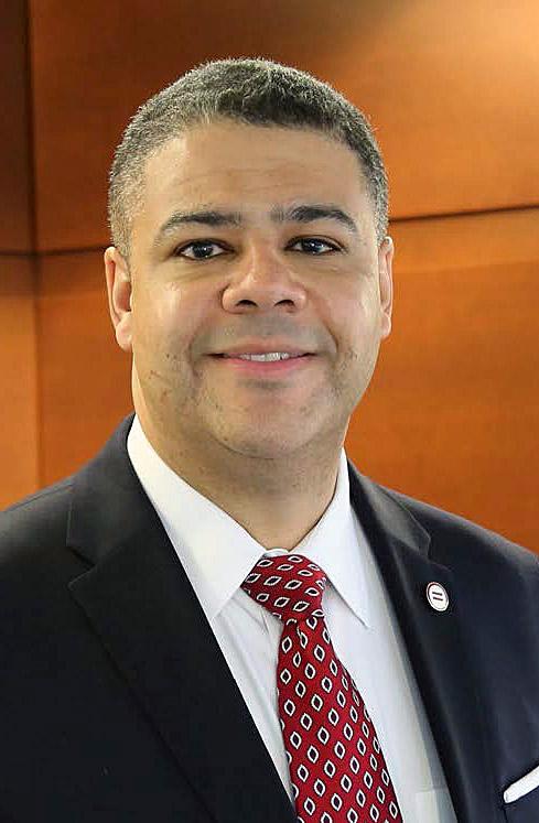 Headshot of man w/short brown hair, dark blazer, white collared shirt, red tie, smiling with wood panels behind