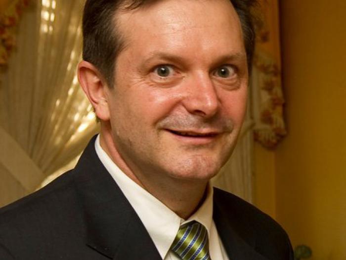 Dean Smith in dark suit, white shirt, shiny moss green tie and brown hair with curtains in the background