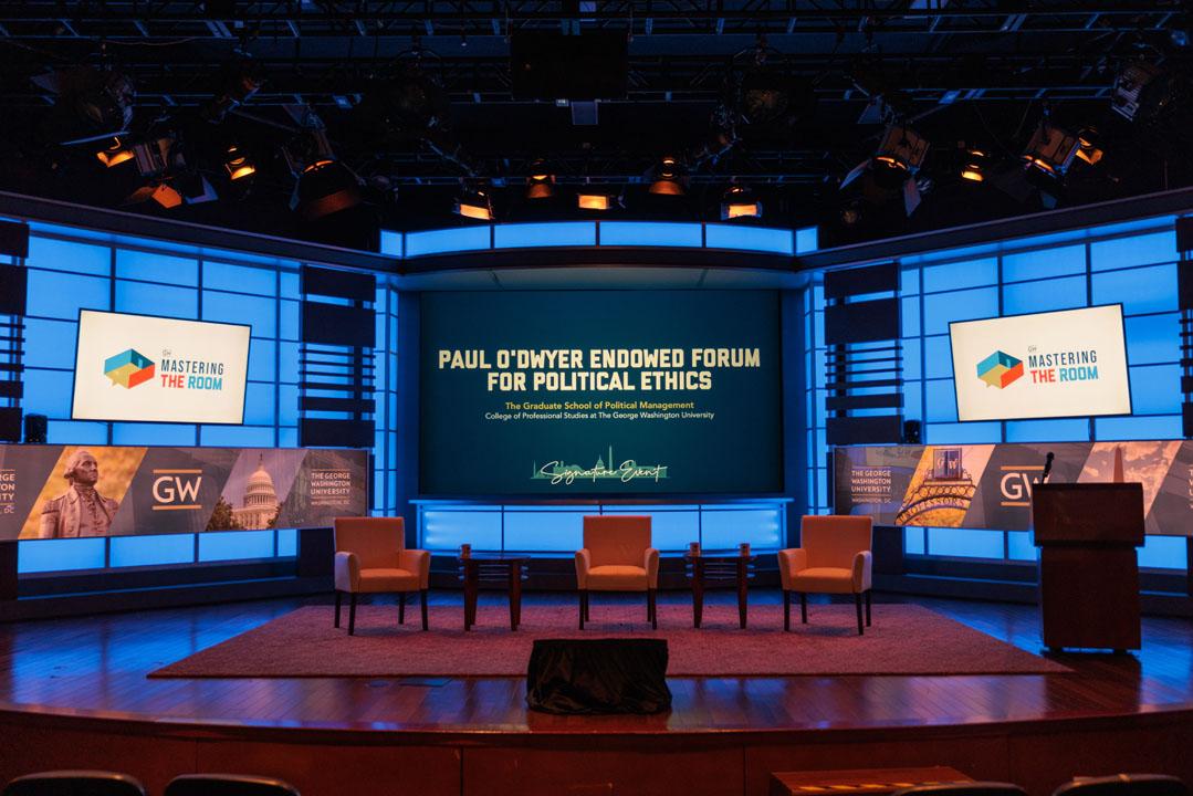 Empty stage with three chairs on it and screens behind them that read "Paul O'Dwyer Endowed Forum for Political Ethics" and "Mastering the Room"